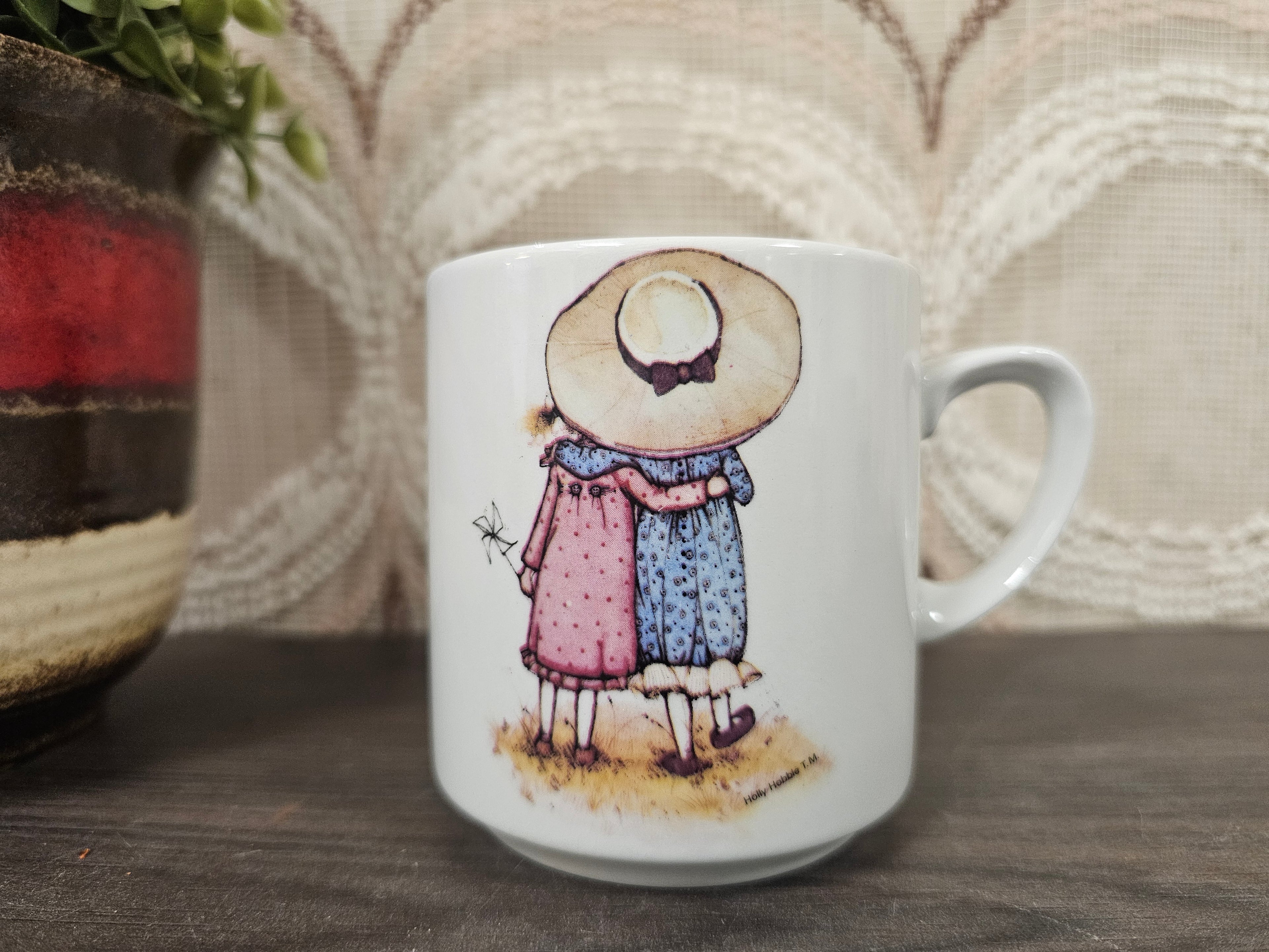 Witte vintage Holly Hobbie beker met tekening van twee meisjes van achteren, één met grote hoed, armen om elkaar heen.