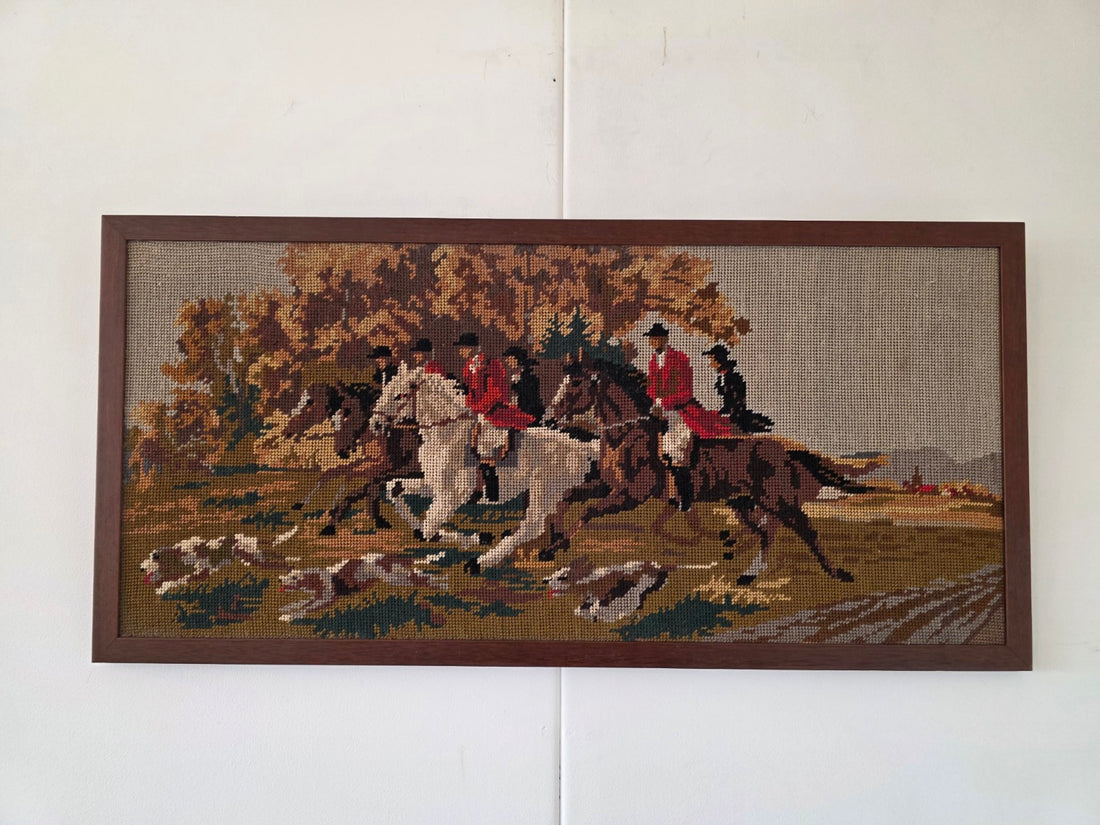 Vintage borduurwerk met ruiters op witte en bruine paarden, jachthonden en herfstbomen in een donkerbruine houten lijst.