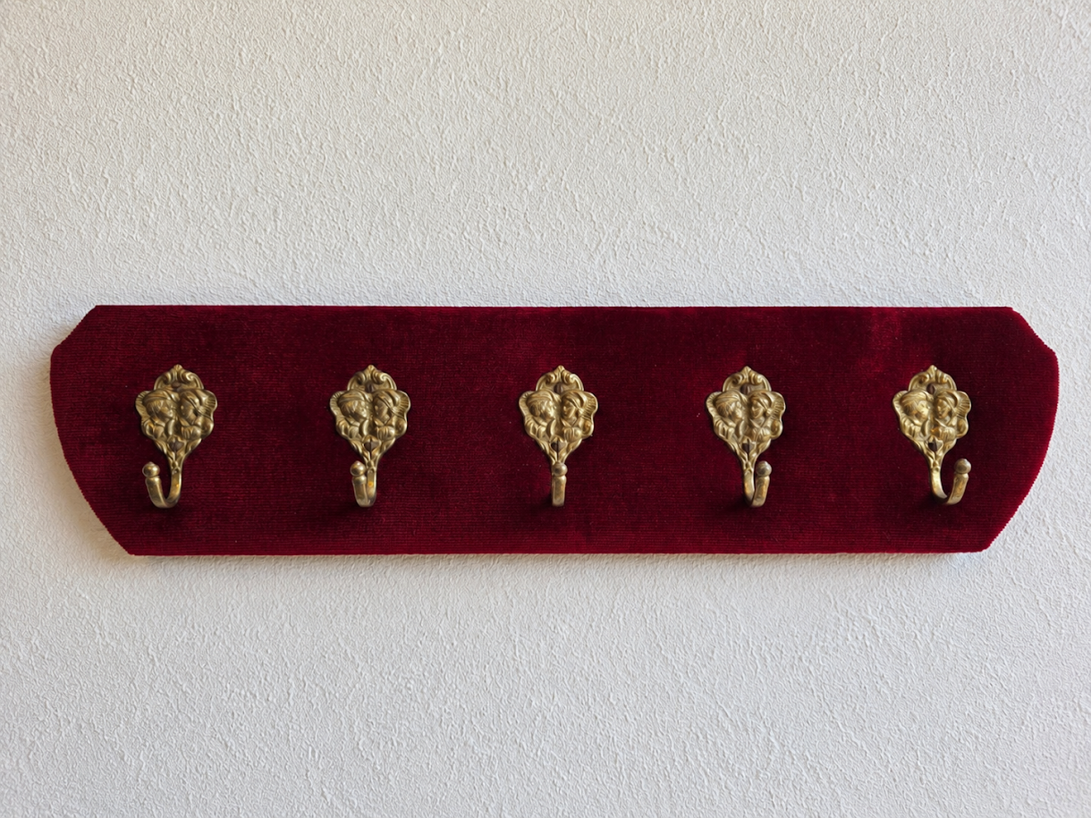 Vintage kapstok met donkerrode corduroy bekleding en vijf messing haken met decoratief gezicht.