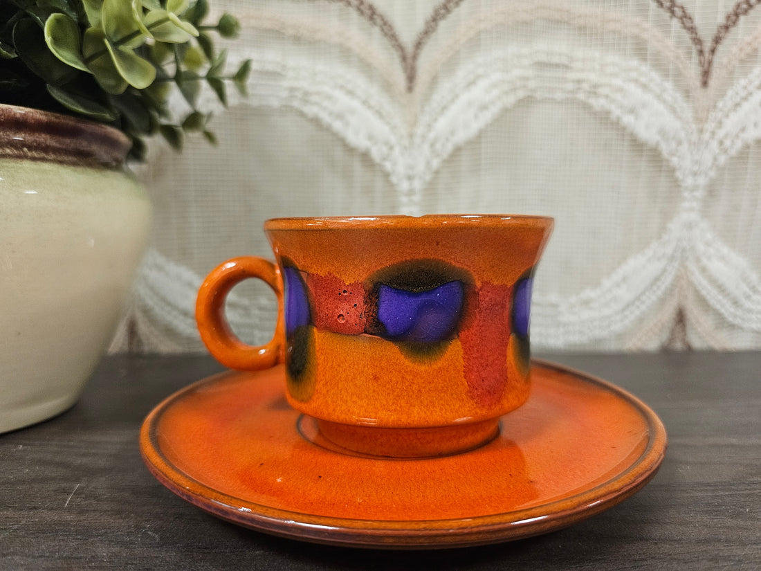 Vintage kop en schotel met oranje glazuur en paars-rood fat lava effect, jaren ’70 stijl, zichtbaar met kleine chipjes.