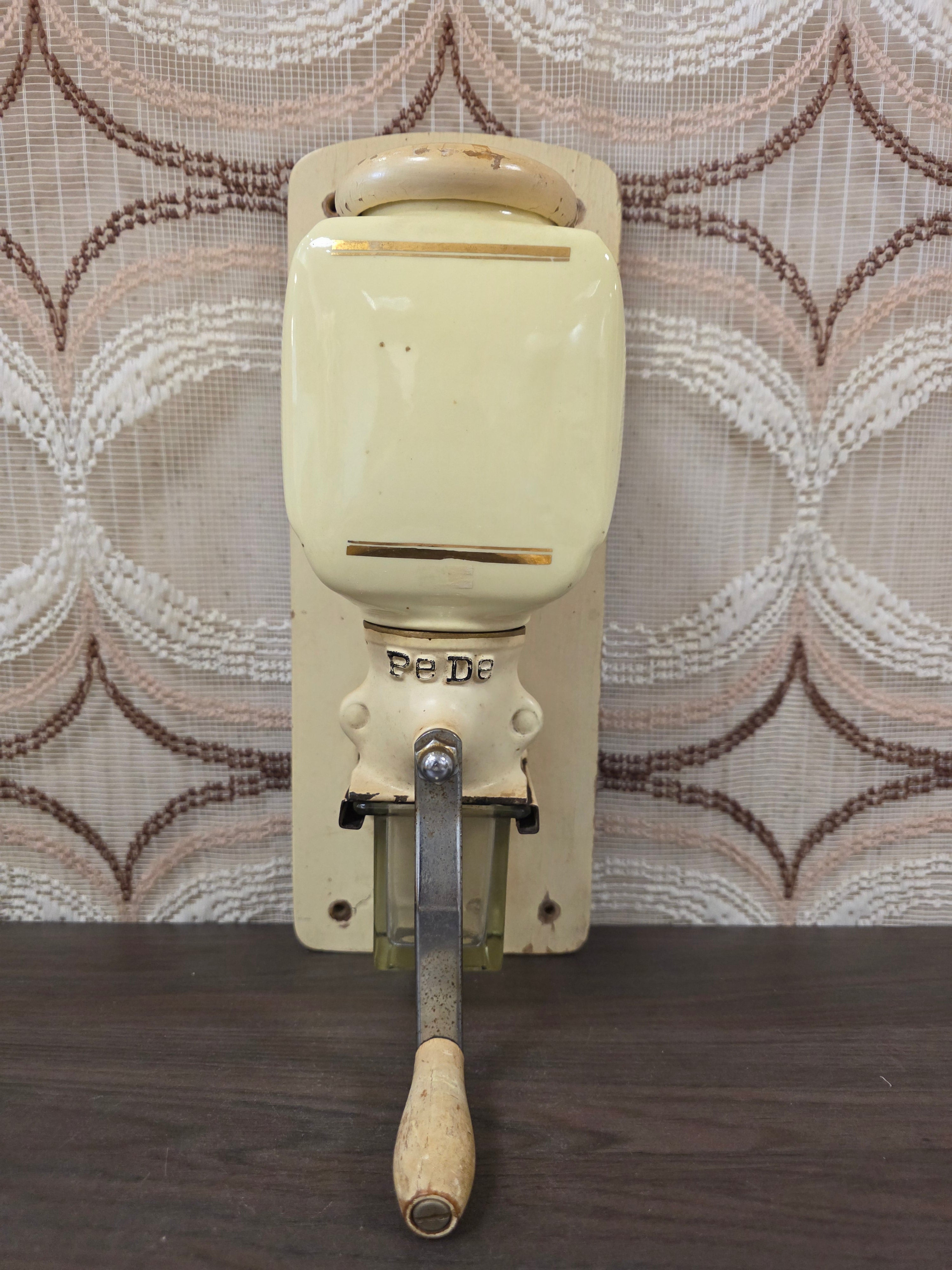 Vintage PeDe wandkoffiemolen in crèmekleur met glazen opvangbakje en houten slingerknop.