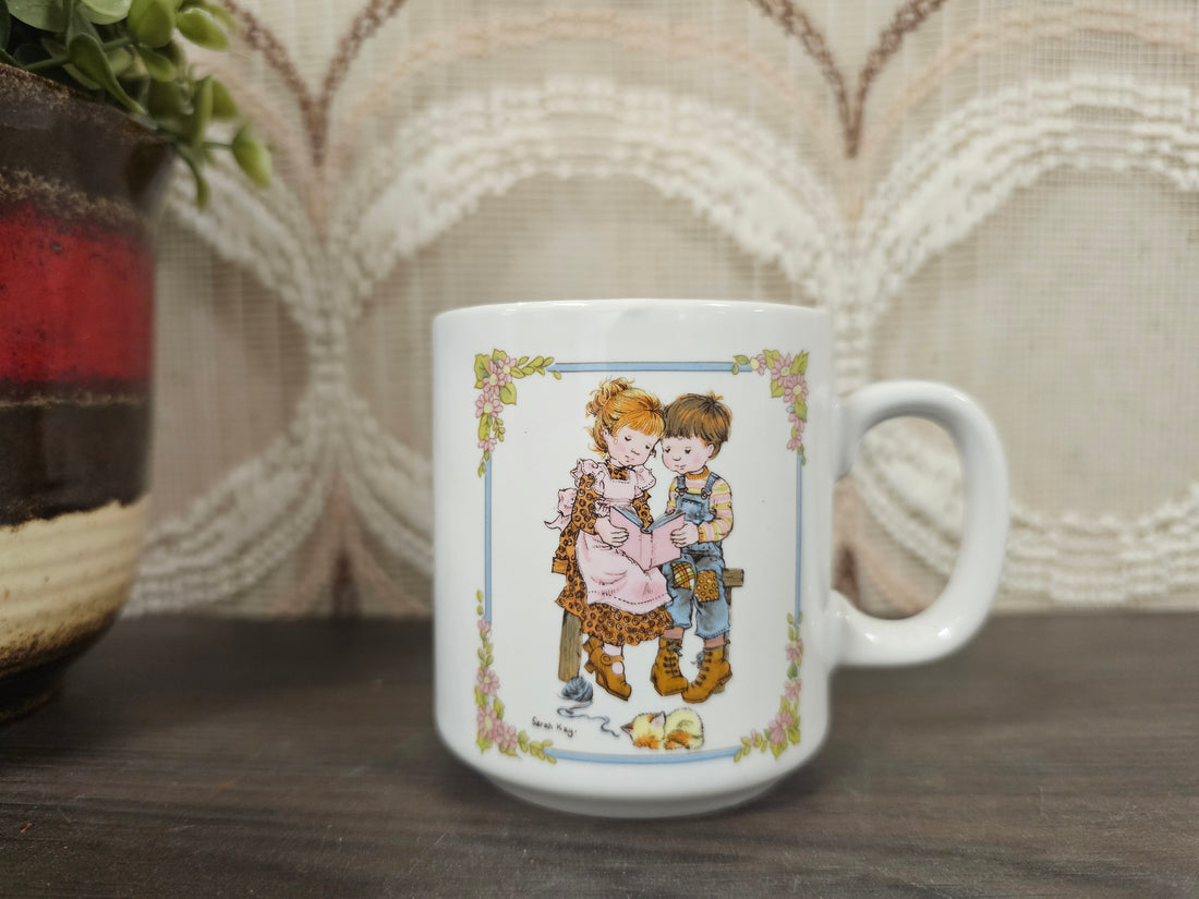 Witte vintage beker met Sarah Kay tekening van twee kinderen op een bankje die samen in een boek kijken.