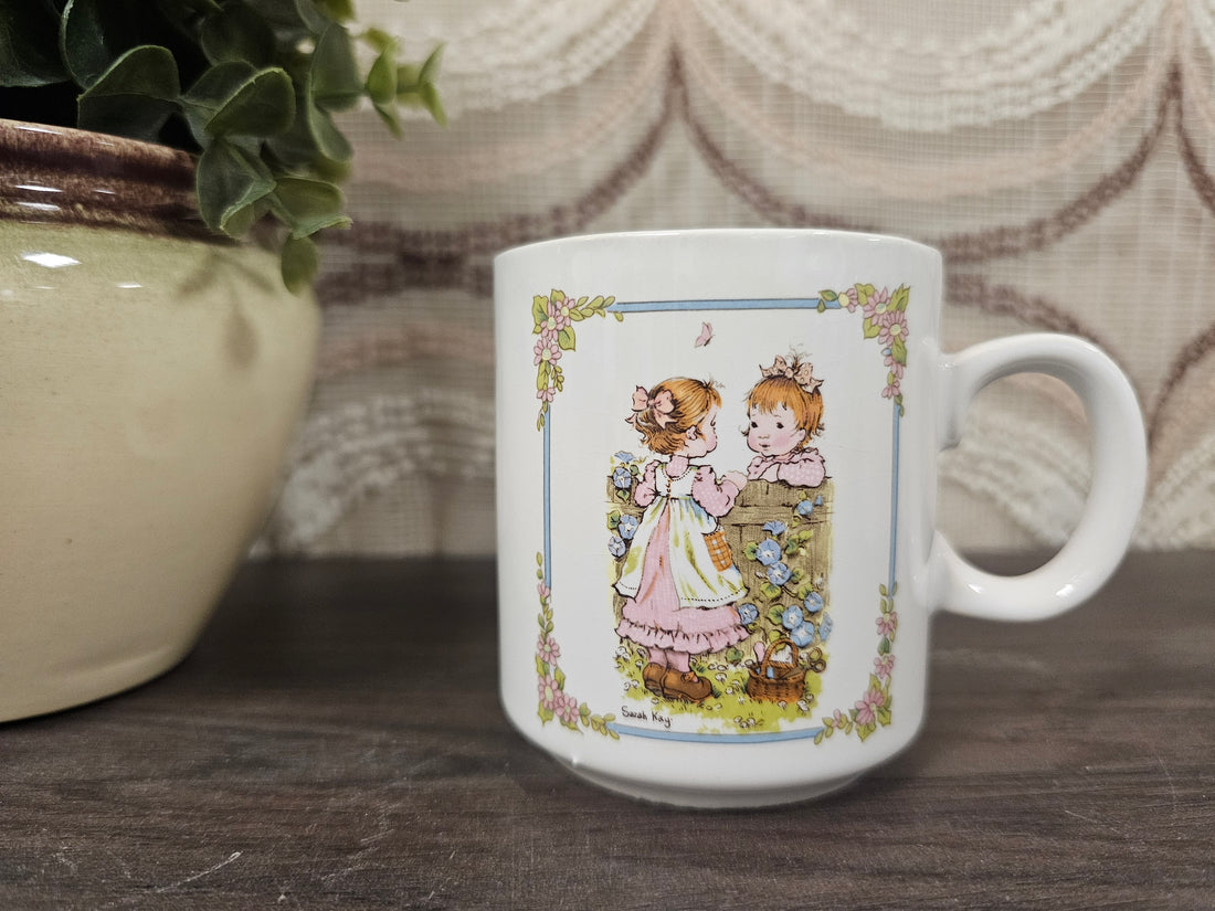 
Vintage Sarah Kay koffiebeker met twee kinderen aan bloemenmuur – wit keramiek
