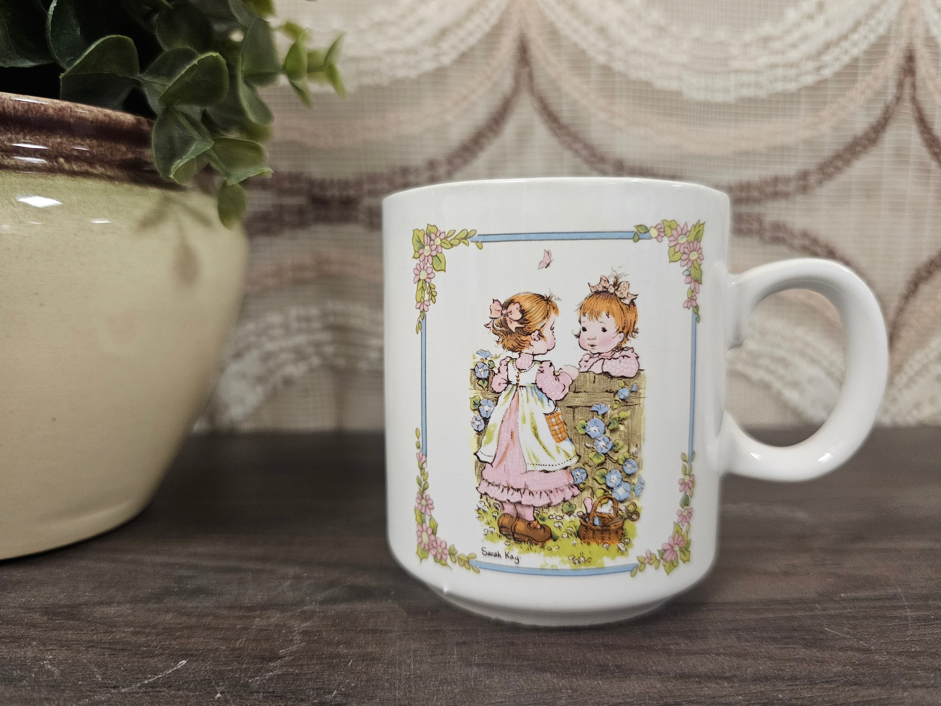 
Vintage Sarah Kay koffiebeker met twee kinderen aan bloemenmuur – wit keramiek