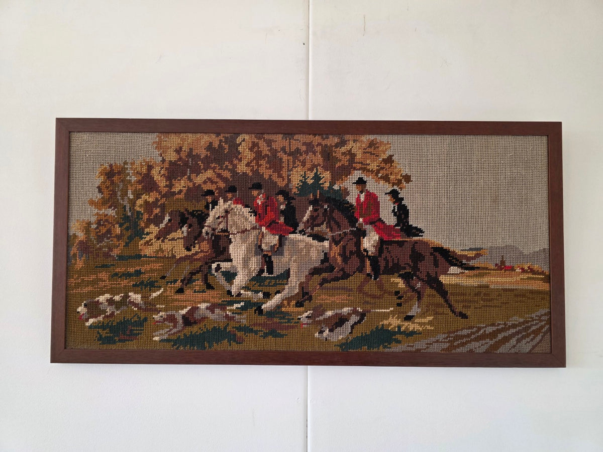 Vintage borduurwerk met ruiters op witte en bruine paarden, jachthonden en herfstbomen in een donkerbruine houten lijst.