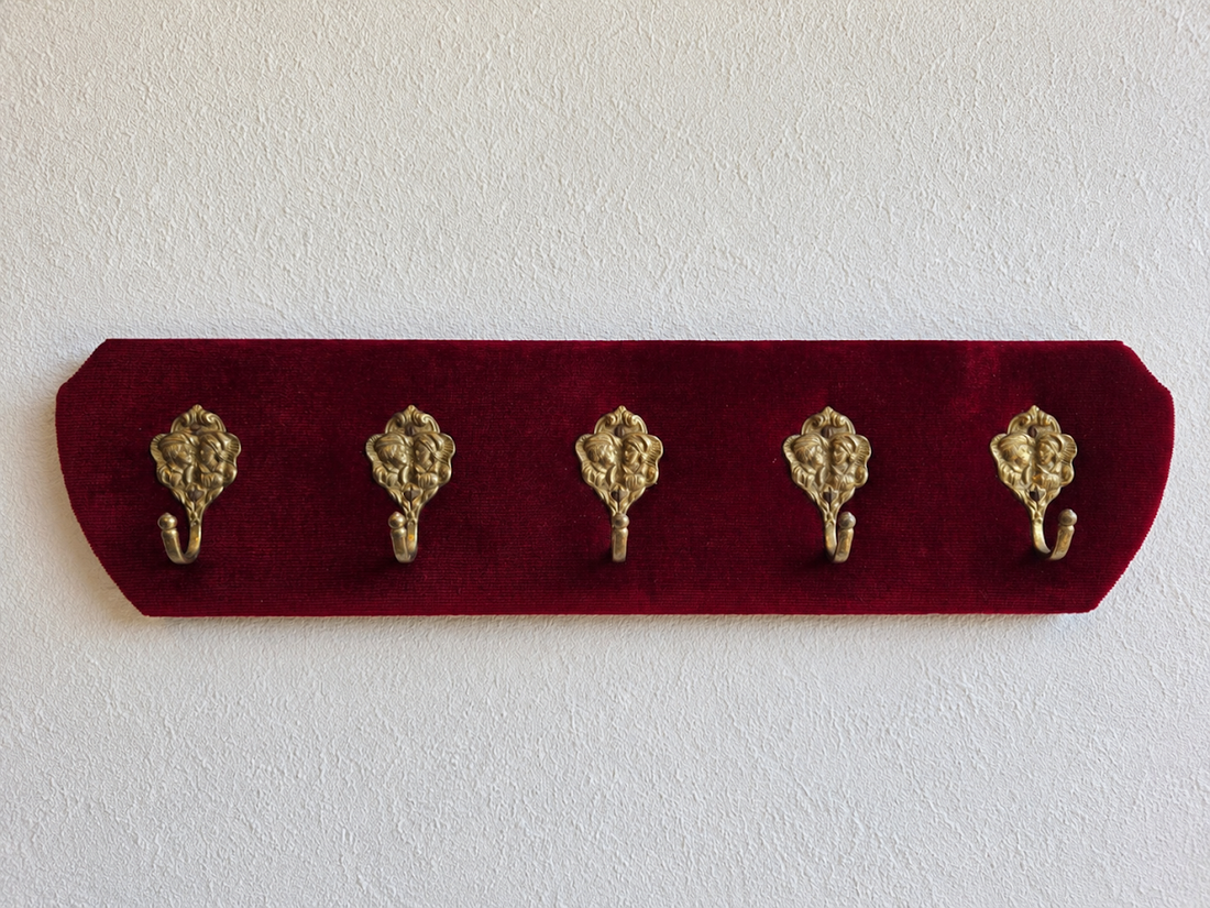 Vintage kapstok met donkerrode corduroy bekleding en vijf messing haken met decoratief gezicht.